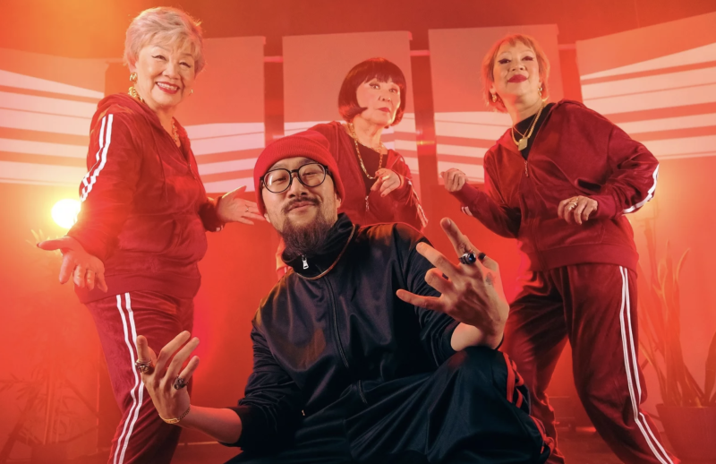 A young Asian man crouches on the floor wearing track suit and red beanie, with a proud expression. Behind him three senior Asian women pose in red Adidas track suits.