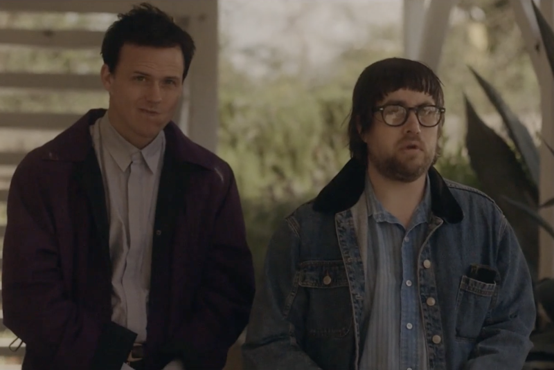An incredulous-looking man wearing a button up shirt and purple raincoat stands next to a confused-looking man with bowl haircut and black spectacles, wearing a blue denim shirt and matching jacket.