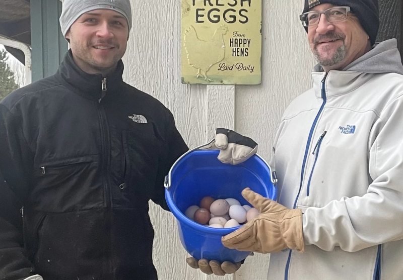 Two men, warmly dressed, hold a blue bucket that’s about half full of multi-colored eggs.