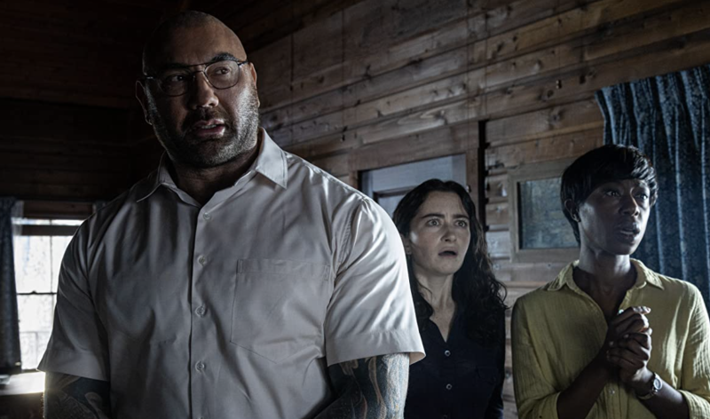 A very large muscular man wearing a white, short-sleeved shirt and spectacles stands inside a wooden cabin. Behind him is a white woman with dark brown hair and a Black woman wearing a yellow shirt. They all look distracted and concerned.