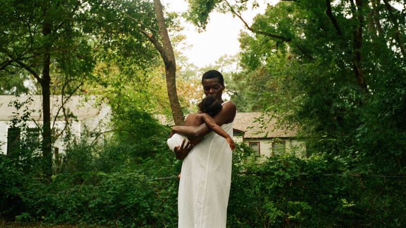 A slender Black woman wearing a simple long white smock, cradles her baby closely in her arms. She is standing outside with lush green trees and a hedge behind her.