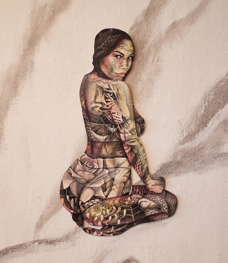 a wooden canvas in the shape of a woman kneeling down and looking over her shoulder assertively. Her skin is covered in tattoos, including a large tiger on her back.