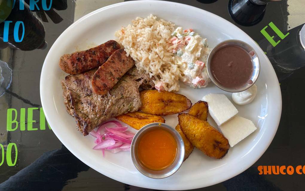 Overhead view of a Guatemalan churrasco chapin plate: steak, rice, potato salad, fried plantains, cheese, and little tubs of salsa and refried beans.