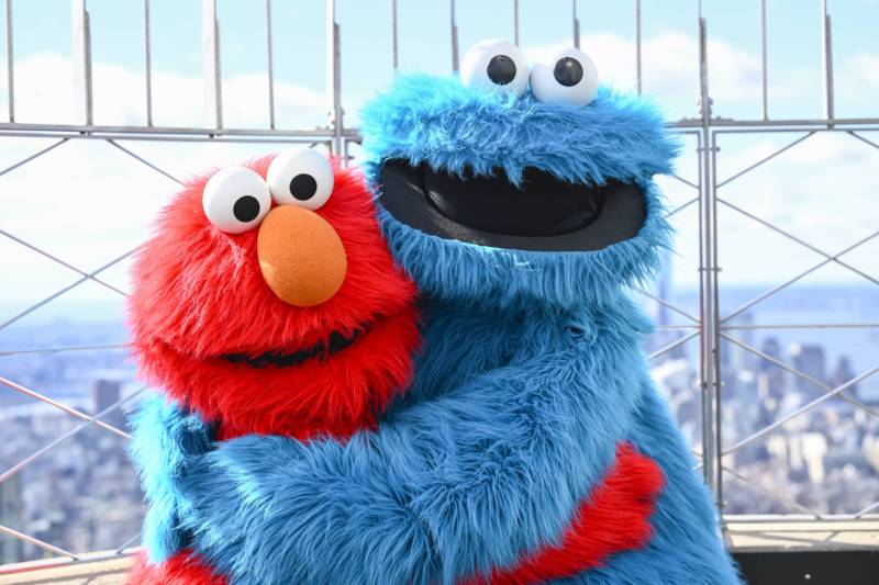 A red furry puppet and a blue fluffy puppet wrap their arms around each other, smiling.