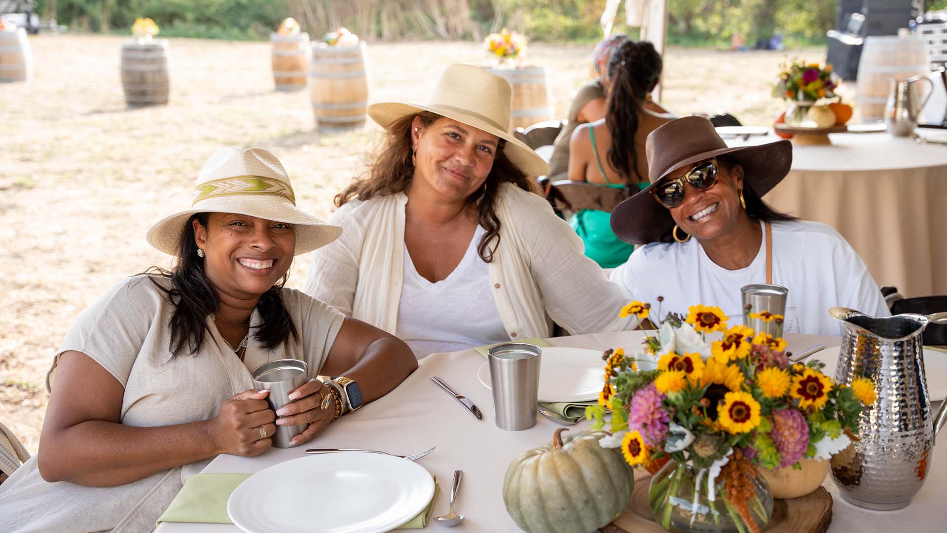 Three smiling women dressed in all white sit at a table outdoors, awaiting the start of a meal.
