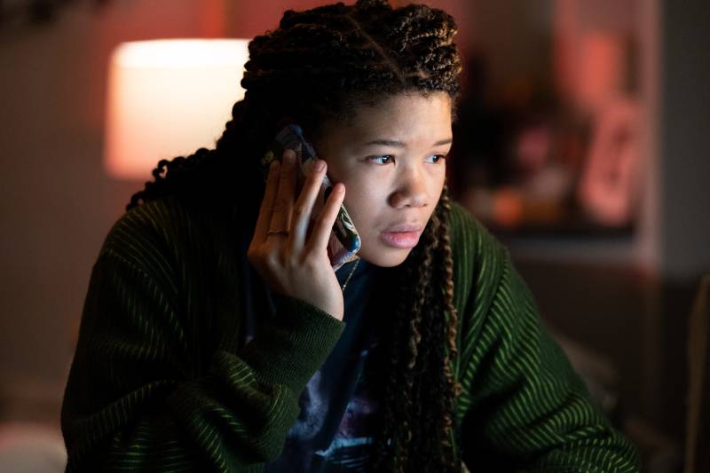 A young Black woman with long braids hovers in front of the light of a computer screen, her cell phone pressed to her ear.