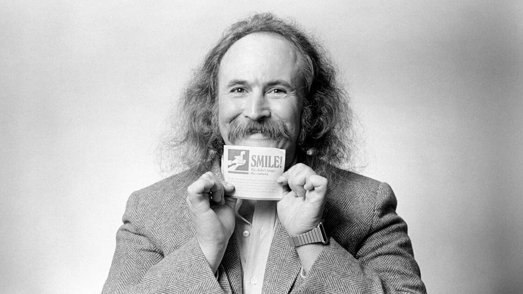 a black and white photo of a middle aged man with long hair and a mustache holding a card that reads 'smile!'