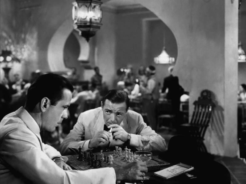 Two men wearing white dinner jackets and black bow ties sit before a chessboard in the corner of a busy restaurant. Both wear expressions of concern.