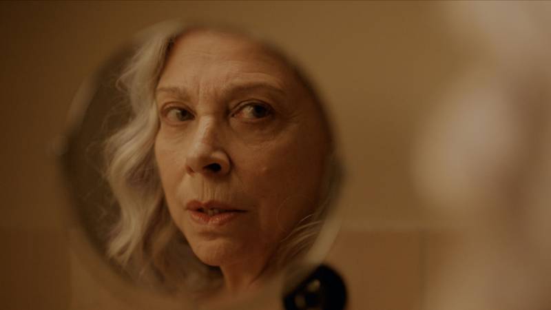 An older woman with pale skin and chin-length grey hair examines her reflection in a small, round bathroom mirror.