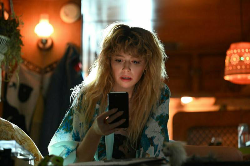 A woman with long, unkempt, blond hair sits in a bohemian looking room, staring at a cell phone screen intently.