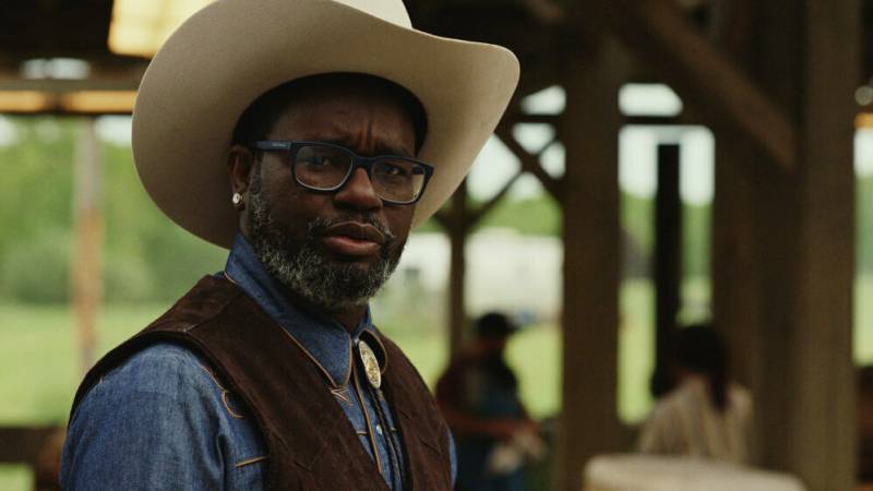 A Black man wearing 1980s-style glasses, a white cowboy hat, blue denim shirt and bolo tie looks confounded.