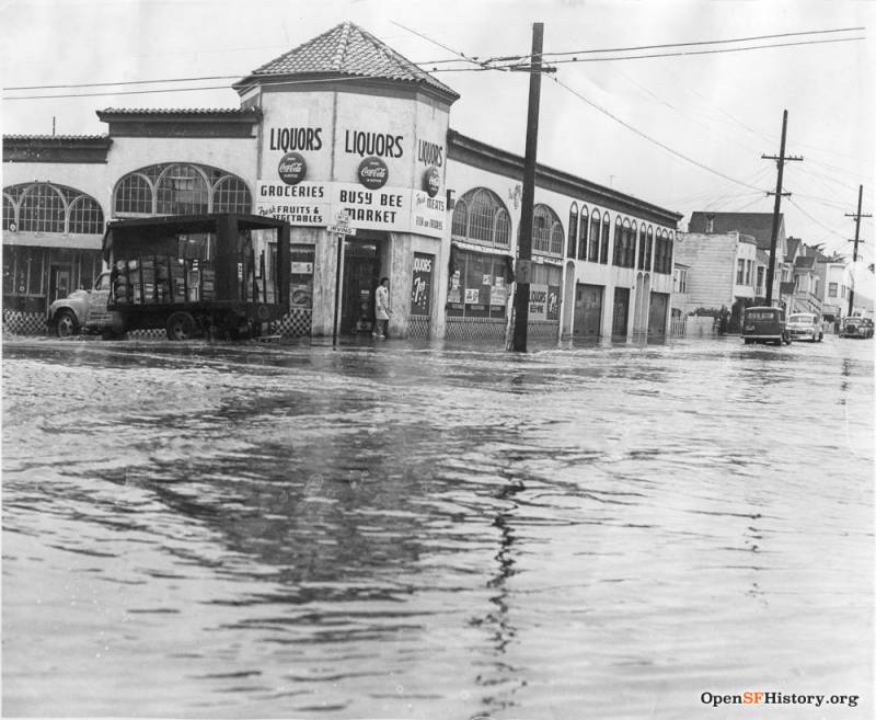 A man stands outside a corner store named Busy Bee Market behind a deserted flooded street.