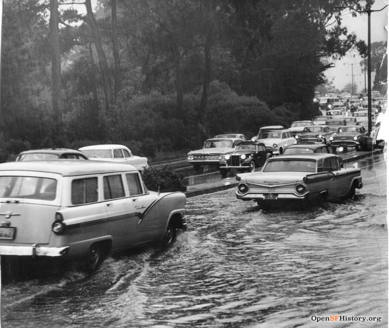 1950s style cars line up in two-way traffic on a flooded, tree-lined road. 
