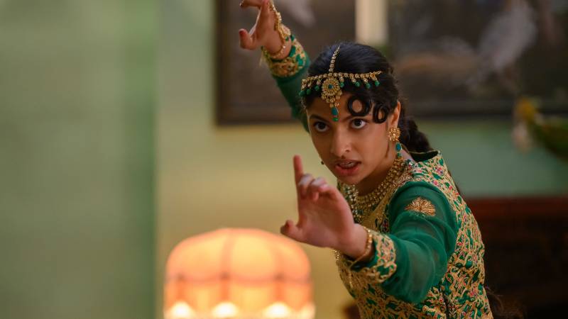 A young South Asian woman wearing a green sari and gold headdress adopts a fighting stance inside a a formally decorated sitting room.