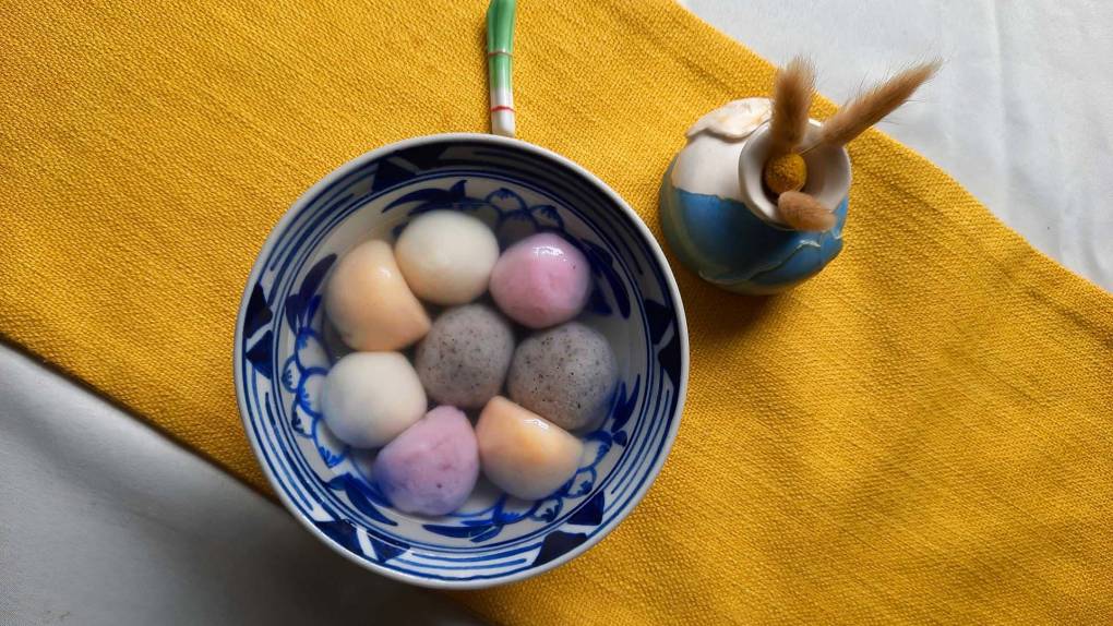 Overhead view of a bowl of colorful glutinous rice balls, or tangyuan, served in a ceramic bowl on gold cloth.