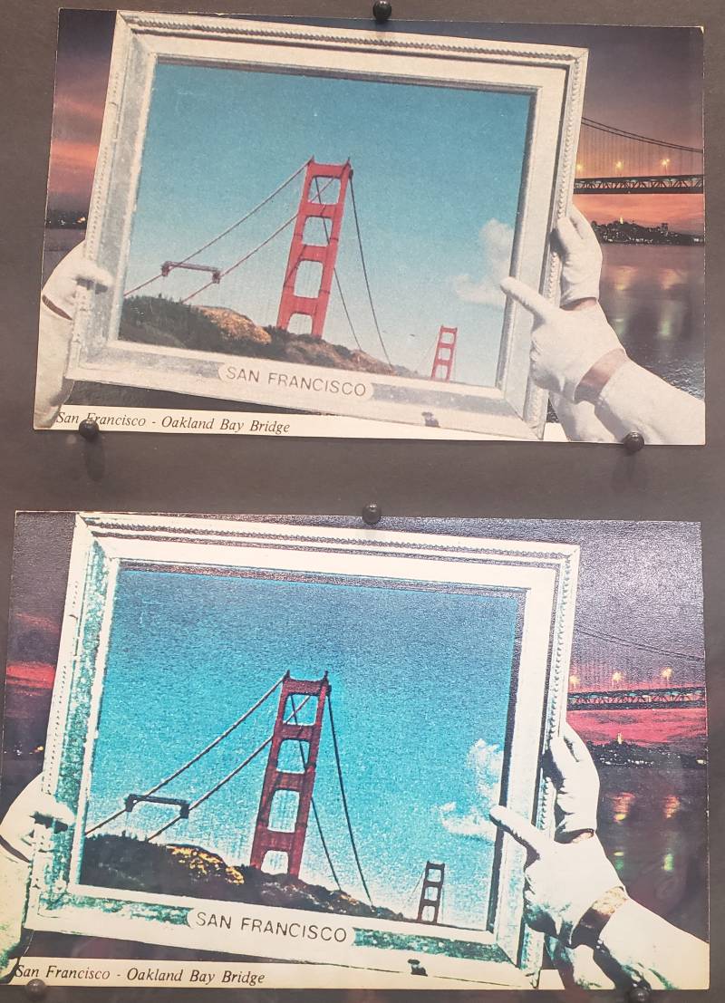 Hands wearing white gloves hold up a framed image of the Golden Gate Bridge over a photograph of the Bay Bridge. The image is duplicated underneath.