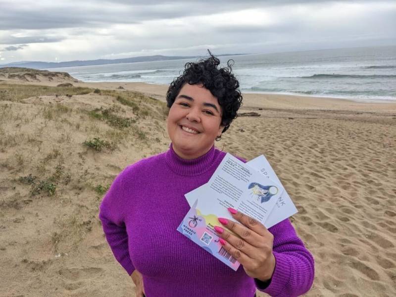 a young Latina presenting woman wears a purple shirt and smiles at the beach while holding her book