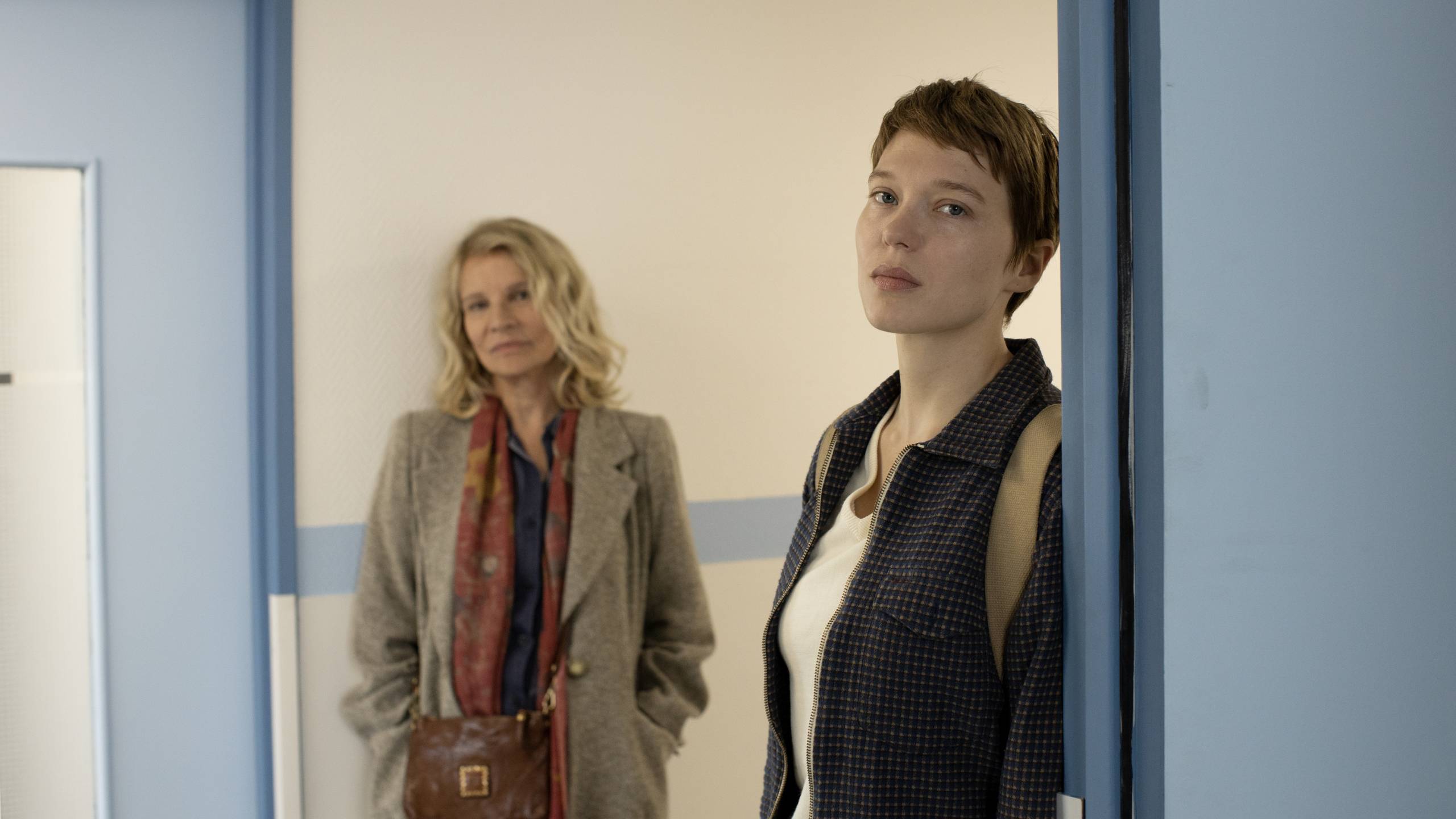 Older white woman leans against institutional wall with younger white woman in doorway