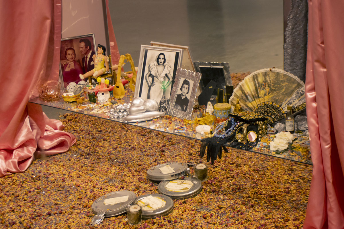 Framed photographs, film canisters and small objects flanked by pink curtains, ground covered in dried flowers