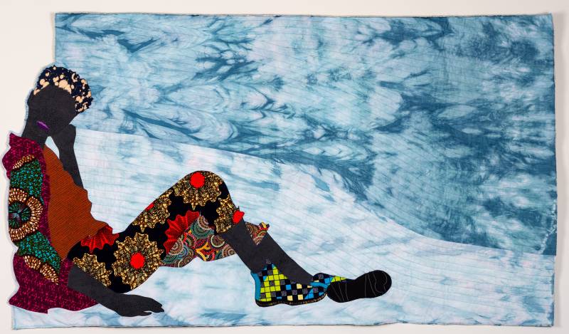 in a textile work, a Black woman in colorful clothing rests sitting on the ground and smiling against a blue backdrop