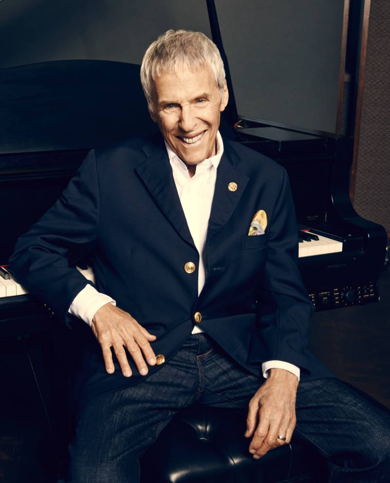 A man sits in front of a piano in a navy suit. He is smiling and facing the camera. 