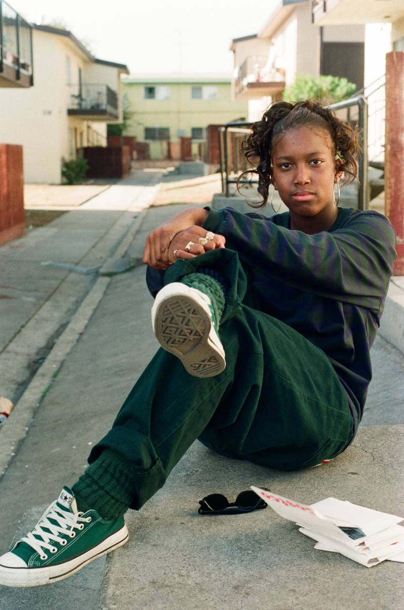 a color photograph shows a young Black woman in green pants and a dark shirt and sneakers sitting on the street looking at the camera