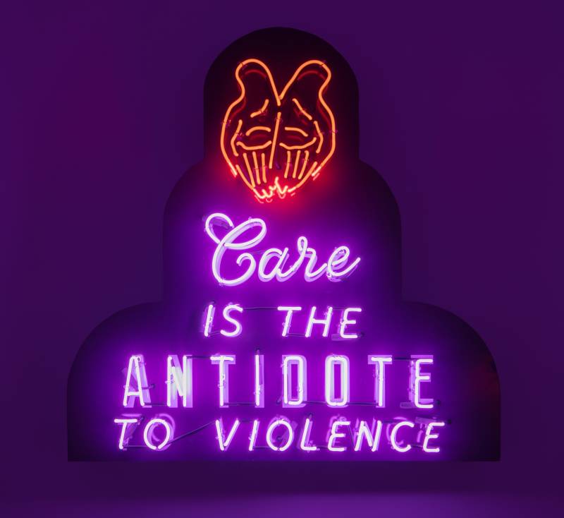 a neon sculpture with a red outline of hands and the words 'care is the antidote to violence' in purple