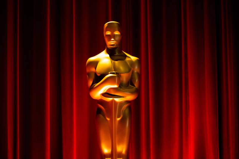 An imposing, life-size gold statue, lit from beneath, stands on stage before closed red curtains.