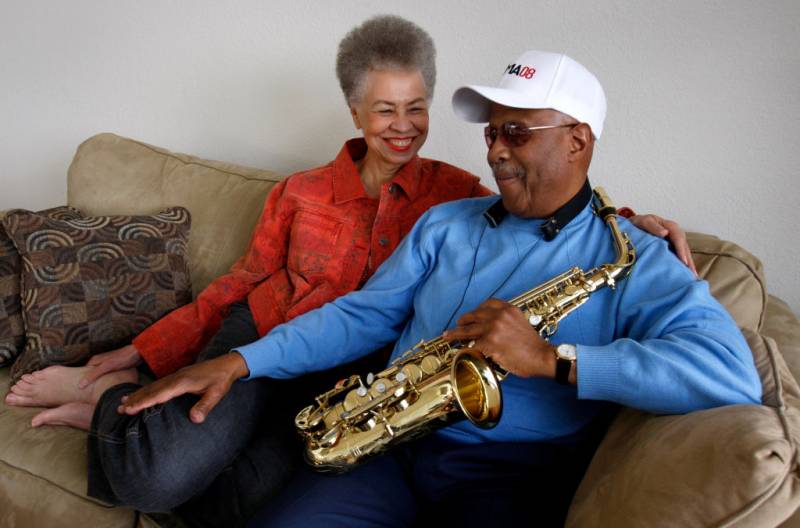 an older Black woman in a red shirt and an older Black man in a blue shirt and white ball cap smile while sitting on a beige couch; he is holding a saxophone and has his hand affectionately on her leg