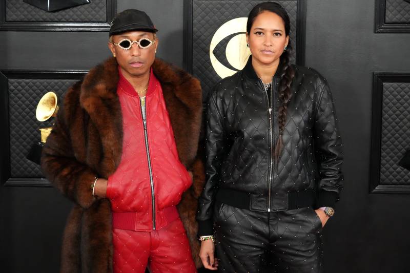 A slender Black man wearing a red leather tracksuit and full length fur coat stands on the red carpet wearing bejeweled sunglasses. At his side is a beautiful mixed race woman with long dark hair. She is wearing a black version of the red tracksuit.