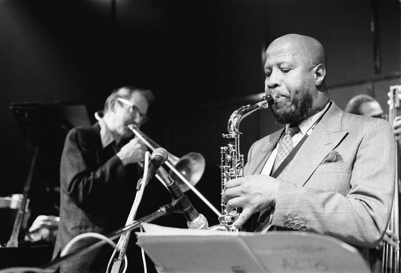 a young Black man in a suit plays the saxophone in the foreground while a white man in a dark suit plays the trumpet behind him in a black and white photo from 1987