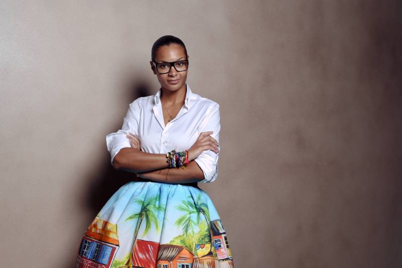 A Black woman with hair pulled back into a tight bun, stands, arms folded, wearing a white shirt tucked into a colorful voluminous skirt depicting a tropical street scene. She is wearing heavy Black rimmed spectacles.