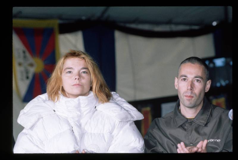 A fresh-faced woman with dirty blond hair sits calmly next to a man with a shaved head wearing a buttoned up shirt. She is wearing a puffy white winter coat.