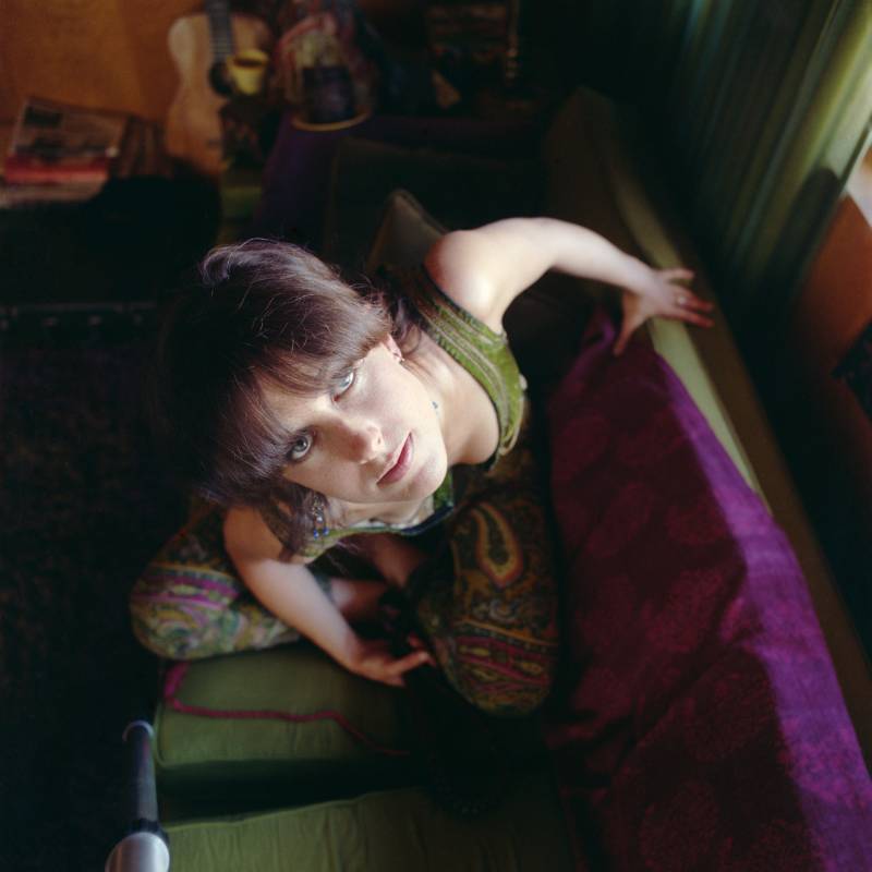 A pretty young woman kneels on a green and purple couch, twisting her head up towards the camera.