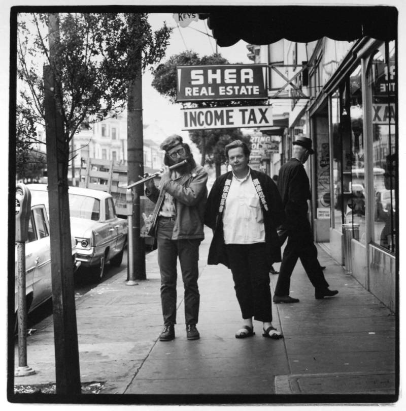 A flute-playing hippie and a bohemian friend, both male, walk along a tree lined street. Behind them a man in a suit and hat walks under a sign that reads 'Sher Real Estate INCOME TAX.’
