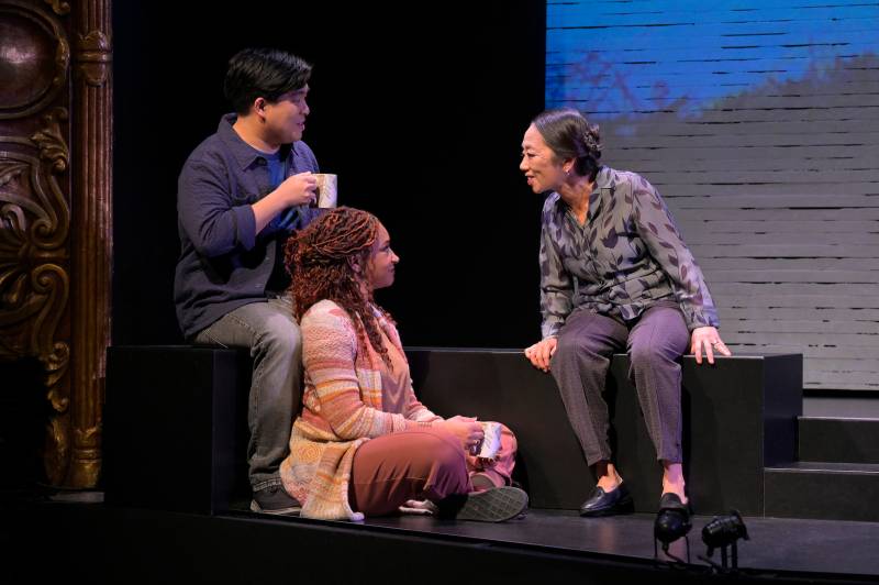 three people on a stage, an Asian man, a Black woman and an older Asian woman, smiling at each other as they sit