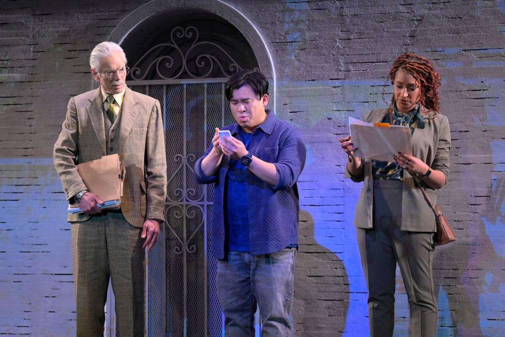 three people stand on a stage in a play -- an older white man, an Asian american younger man and a Black woman. they are looking at phones and pieces of paper