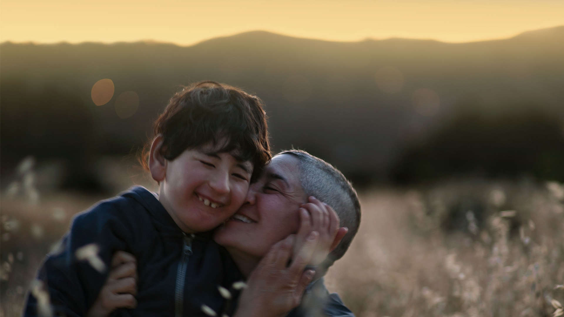 Young boy with dark brown hair smiles and touches face of woman with buzzed hair in golden landscape