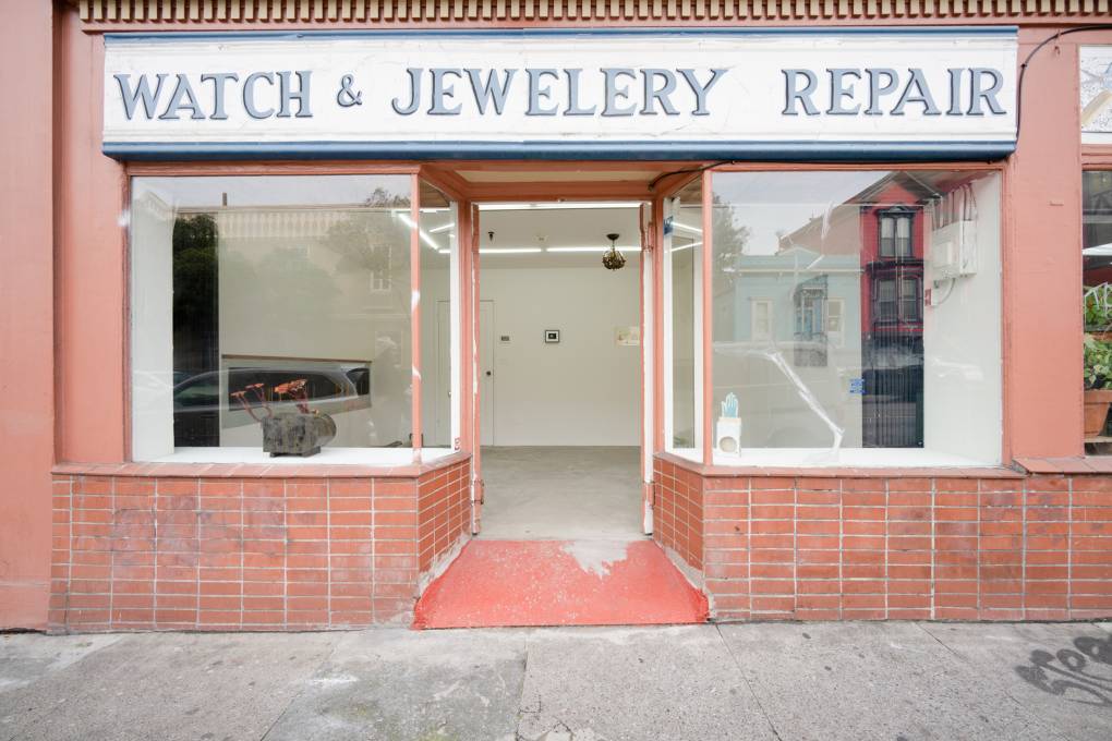 Red bright storefront with white wall gallery inside, sign above door reads 'watch & jewelery repair'