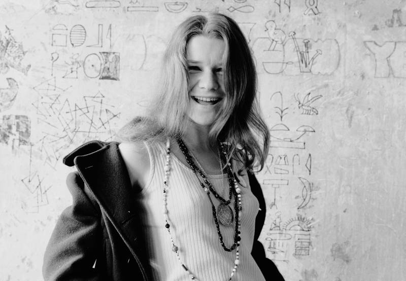 A laughing woman with long brown hair stands before a white wall that's covered in scribbles and messages. She is wearing a simple tank, beaded necklaces and a winter coat that's falling from her shoulders.