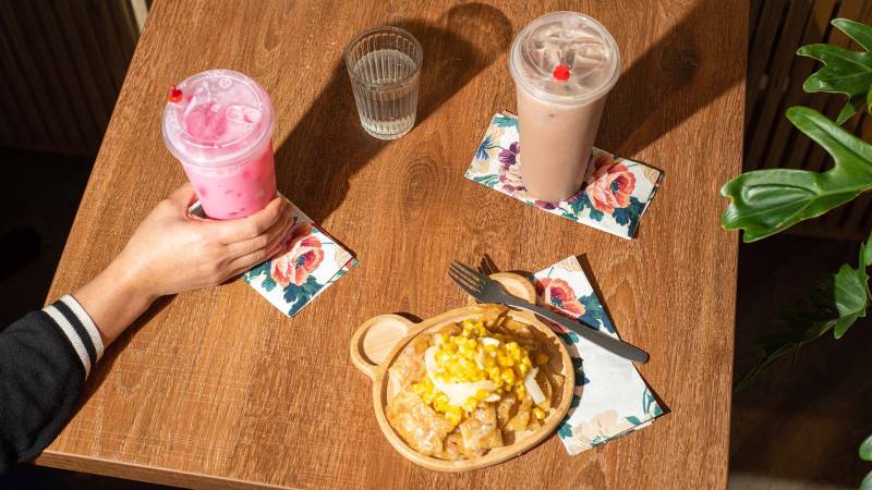 A hand reaches to pick up an icy pink beverage. There's also a iced chocolate drink and a plate of corn roti on the table.