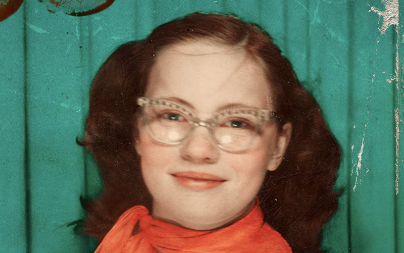 A young woman with red hair pulled back into pigtails smiles sweetly in a photo booth with an orange scarf tied around her neck. She is wearing fashionable 1950s spectacles.