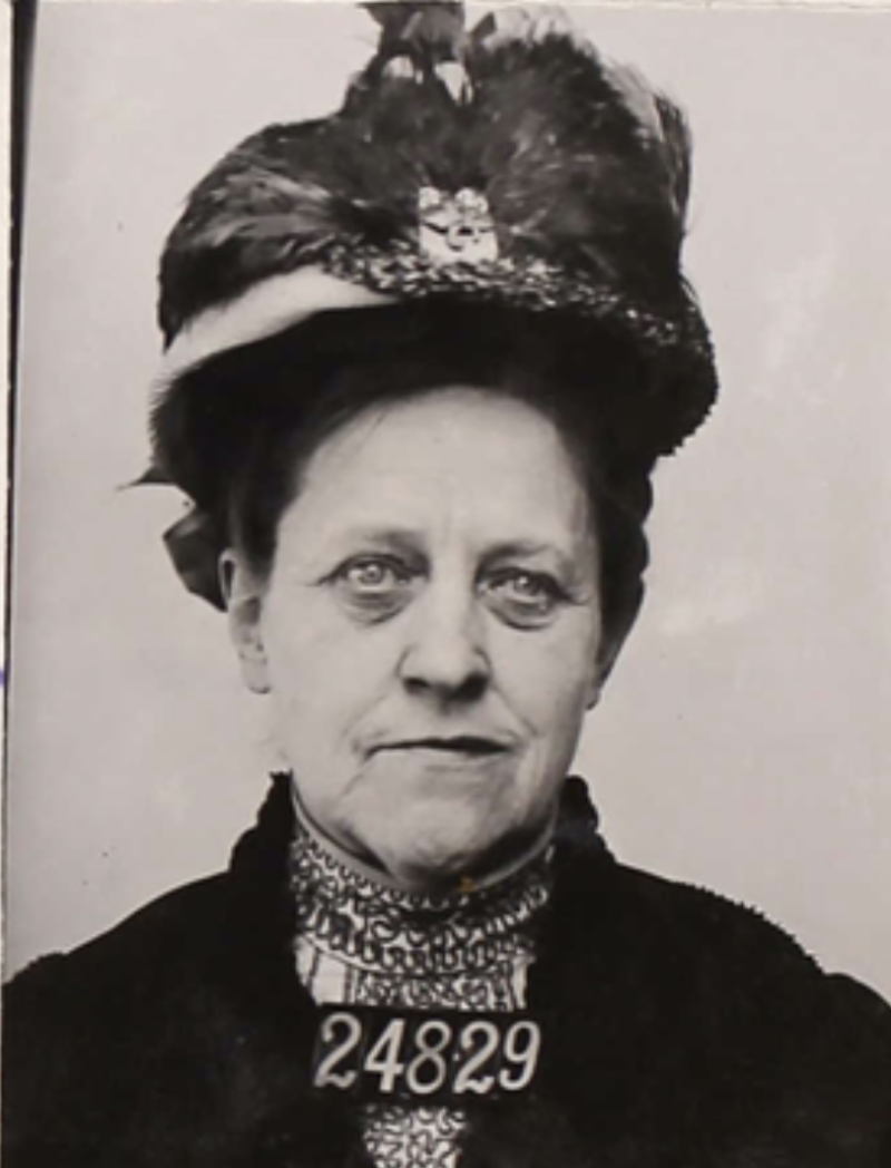 A sour-faced white woman in high collared Victorian clothing and feathered hat stands with a numbered card attached to her chest.