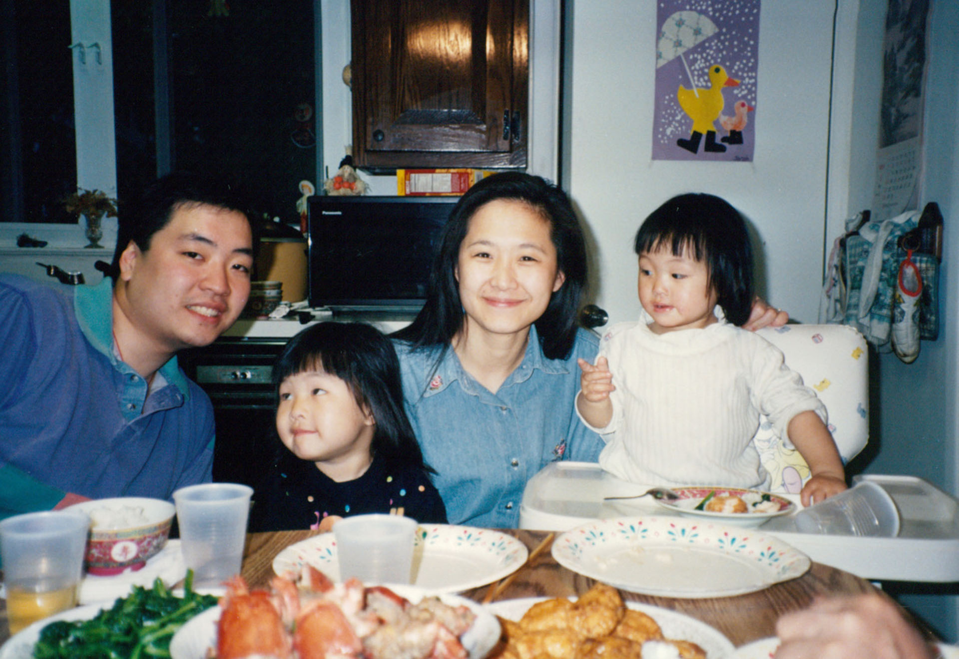 A family photo of a Chinese American family taken during the 1990s. A father, mother and two daughters sit at the dinner table.