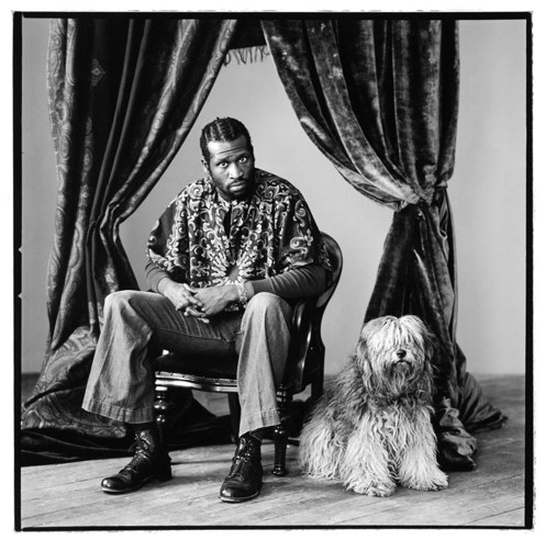 A Black man with cornrows sits in a wooden chair wearing a tie-dye shirt and slacks. He is leaning forward as if in conversation. At his side is a shaggy white dog.