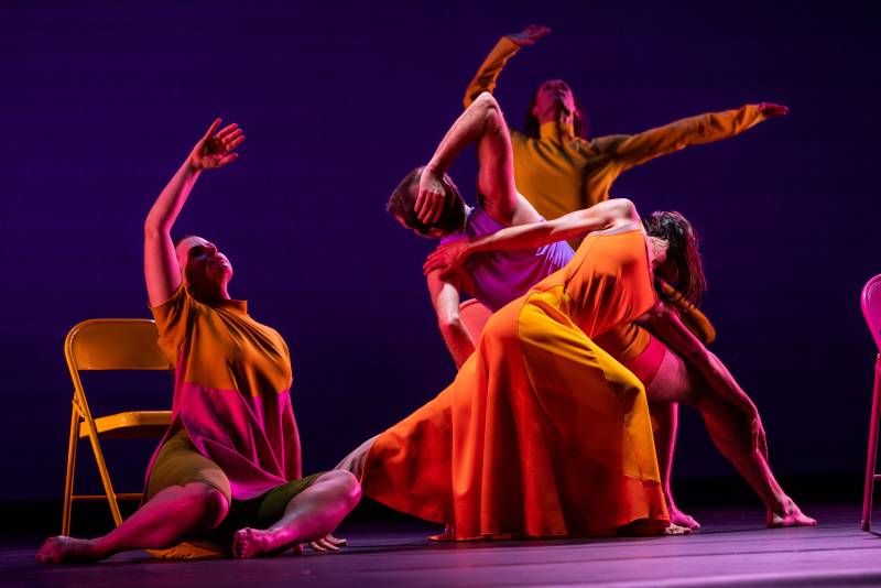 Four dancers are on stage in orange against a backdrop of blue. The photo captures them all mid-motion, but all doing separate movements. One sits on the floor with their arm reaching into the air. Two in the middle are lunging to opposite sides, their torsos crossing with their backs to each other. In the background a fourth sways their arms above themselves.