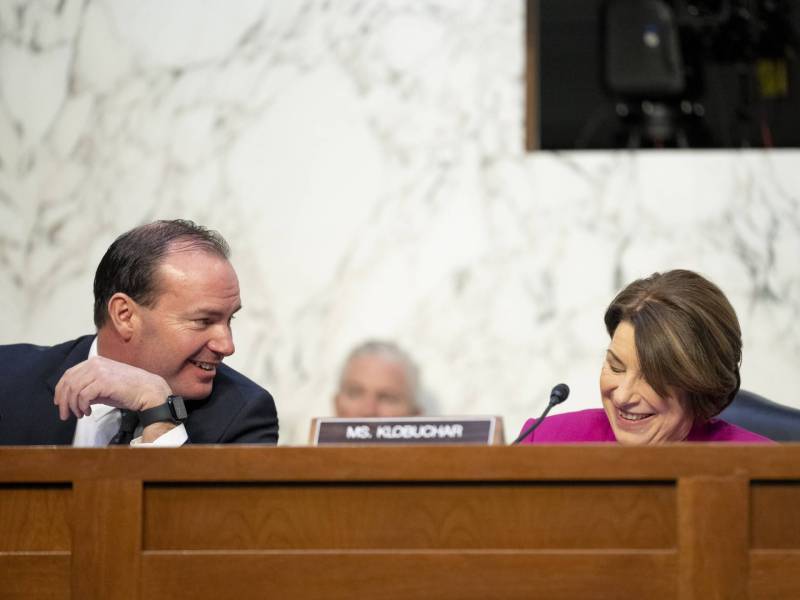 A man with a receding hairline smiles and leans to the right towards a smiling woman wearing a pink suit. She has a small microphone in front of her and a sign that reads 'Ms. Klobuchar.'