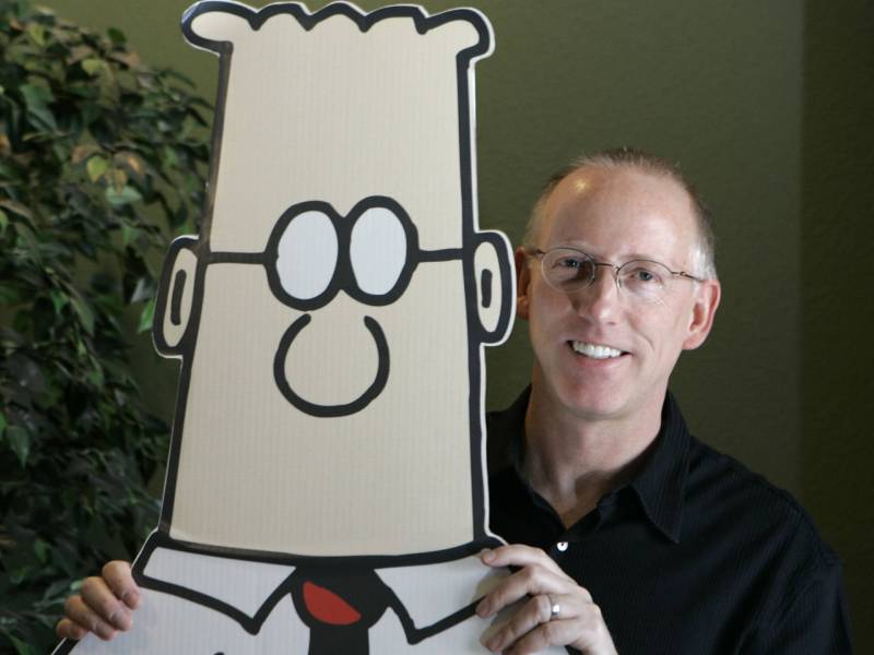A smiling white man with bald head and glasses stands behind a life-size cardboard cutout of a cartoon character wearing red tie and spectacles.