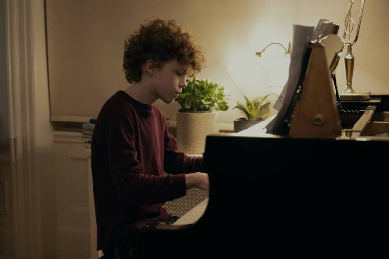A young boy with short, curly hair, sits at a piano. House plants are visible behind him.