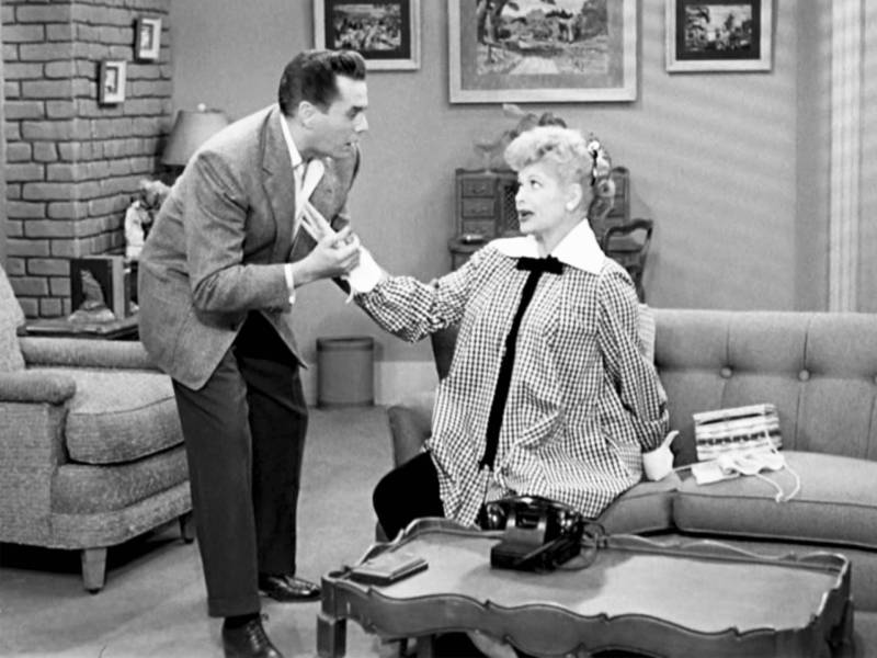 A black and white image of a suave man in 1950s-era suit, holding the hand of a pregnant woman sitting on a couch. She is wearing a billowing pregnancy shirt. 
